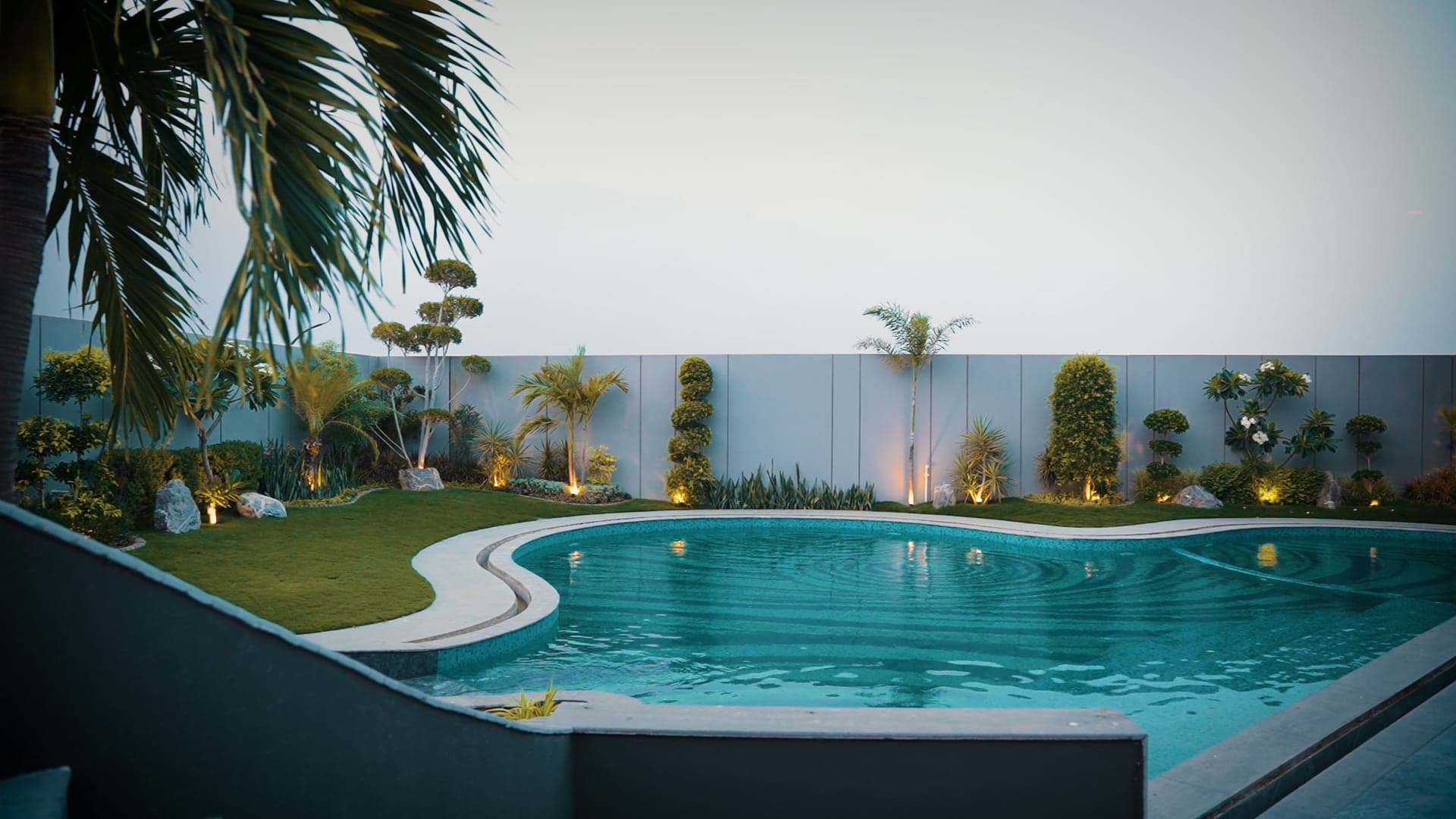 Free-form swimming pool beside a landscaped garden of palms and topiary along an uplit perimeter wall at dusk at FHU Resorts Karachi