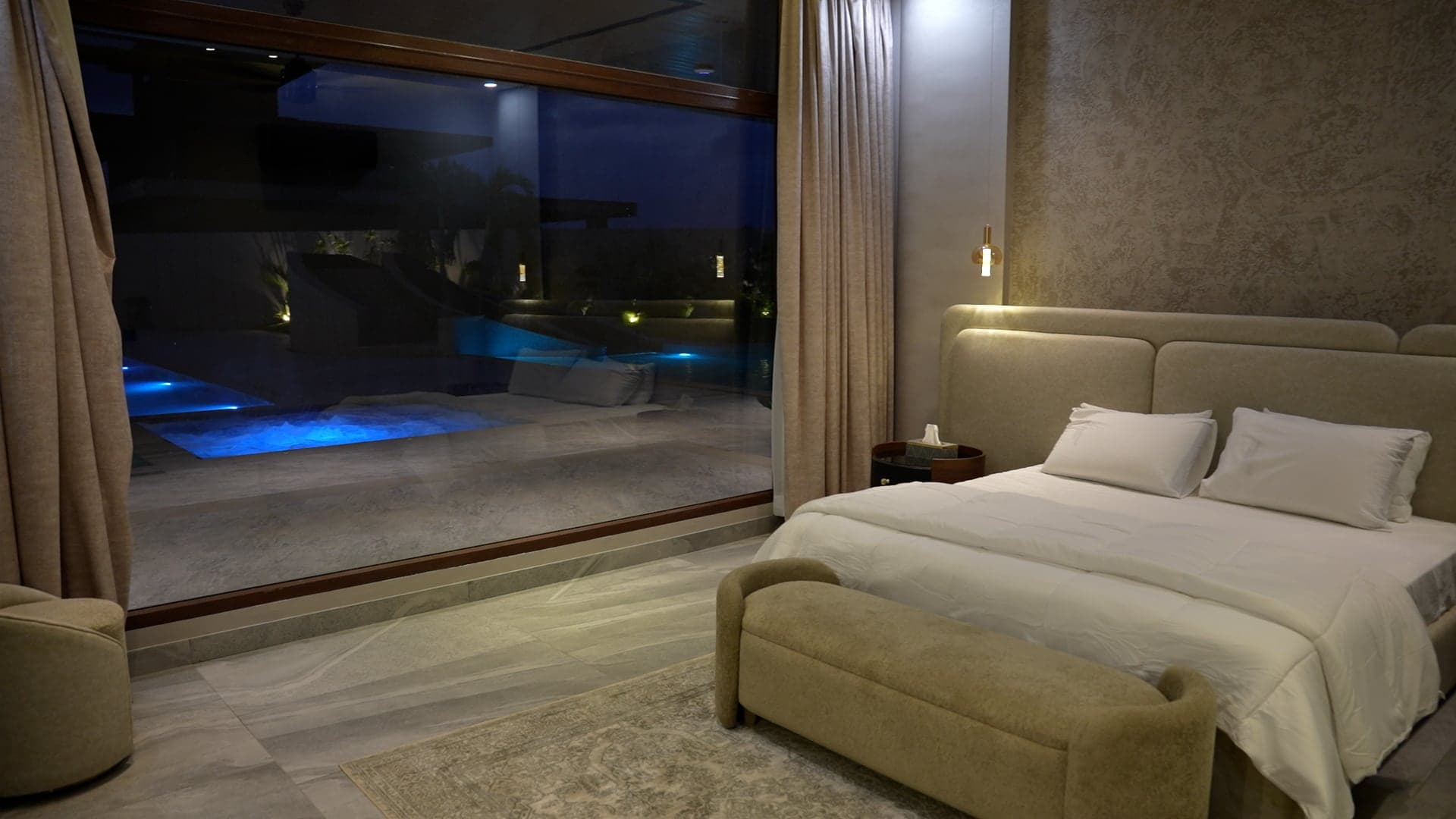 Master bedroom with a floor-to-ceiling picture window overlooking the blue-lit pool at night at FHU Resorts Karachi